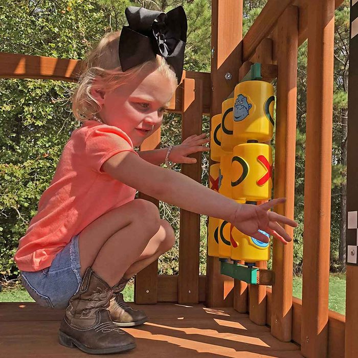 A young girl in a bright shirt plays with colorful, interactive plastic tubes on a wooden playground, surrounded by trees and natural sunlight.
