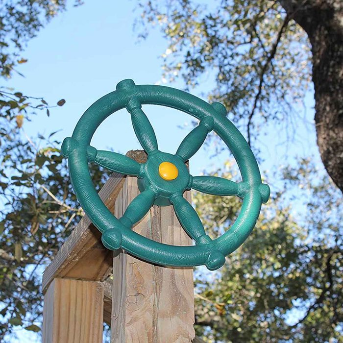 A green steering wheel toy is mounted on a wooden post, surrounded by trees and a clear blue sky.