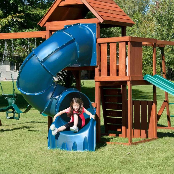 A girl slides down a blue tube slide, enjoying her descent from a wooden playground structure, surrounded by green grass and trees in bright sunlight.