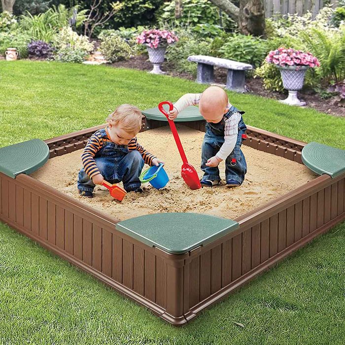 Two children play in a sandbox, scooping sand with colorful toys, surrounded by a lush garden with flowers and greenery in the background.