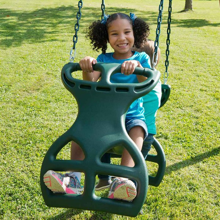 A child with curly hair swings joyfully on a green swing in a grassy park, smiling and holding onto the swing's handles.