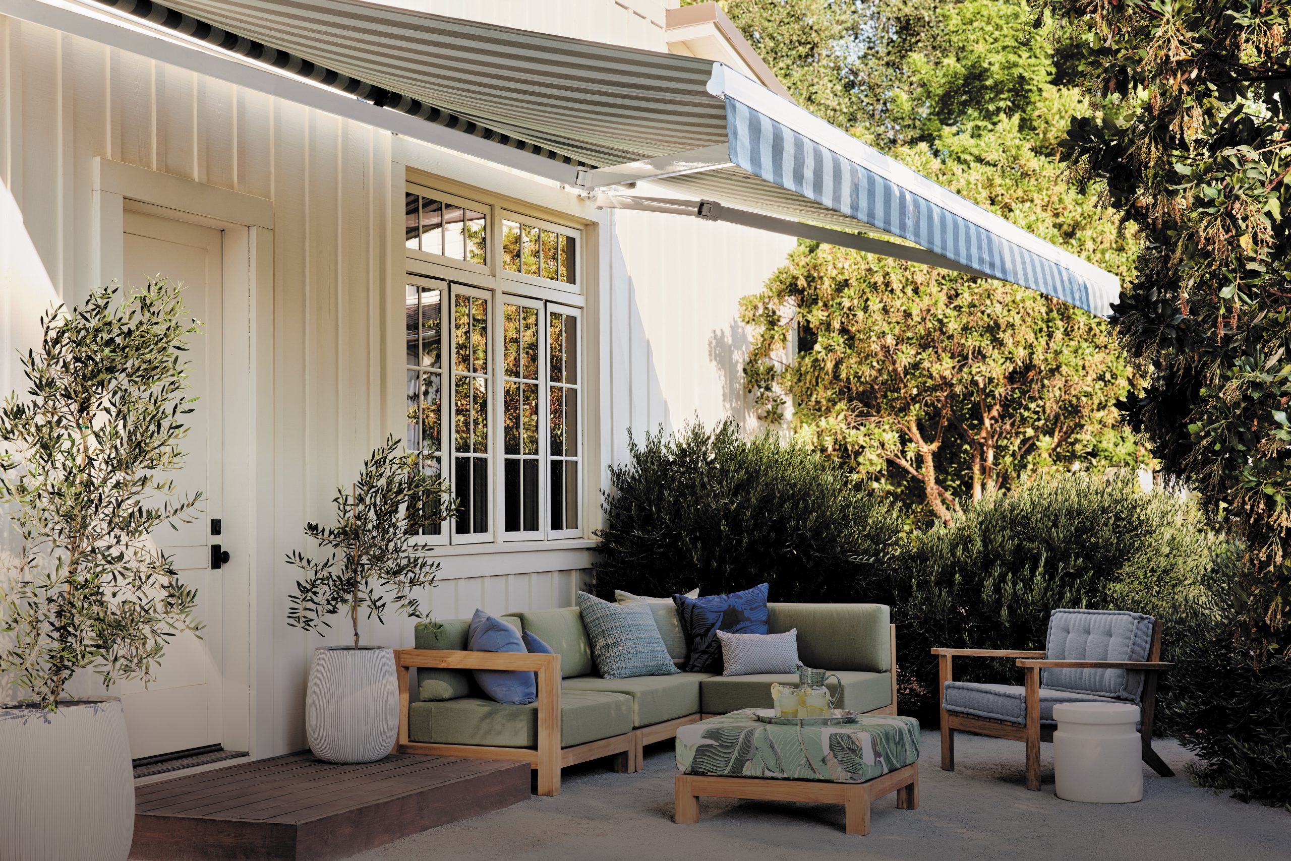 A green couch, adorned with pillows, sits on a patio; an awning extends overhead, surrounded by potted plants and lush greenery.