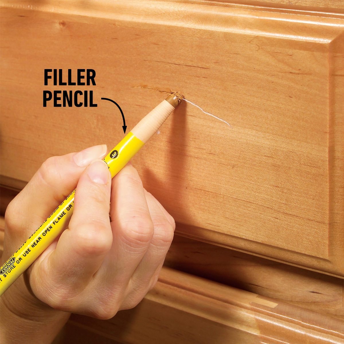 A hand holds a yellow filler pencil, applying it to a small scratch on a wooden surface. The environment suggests a home or furniture setting.