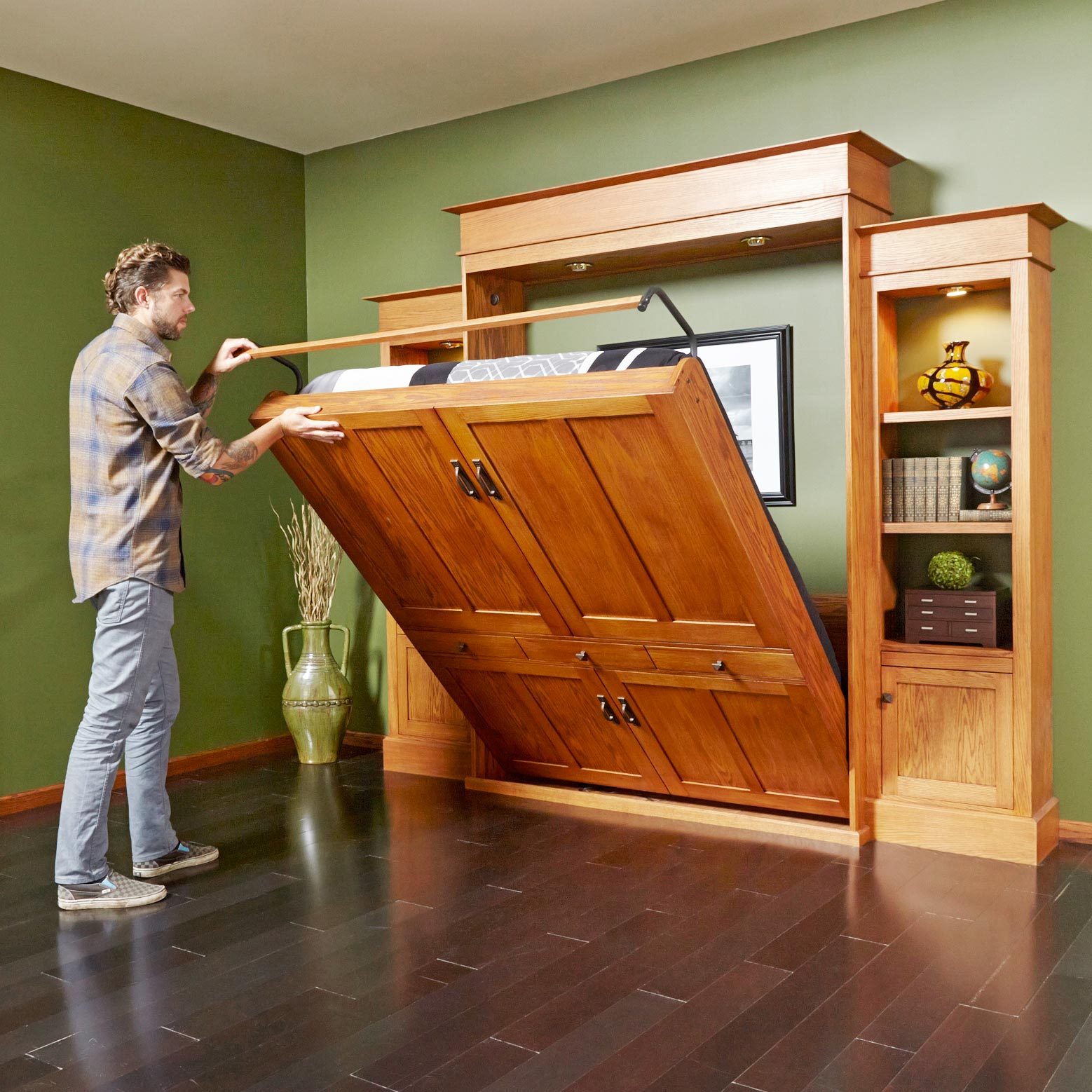 A man raises a murphy bed from a wooden cabinet in a neatly decorated room with green walls and wooden flooring, preparing for bed.