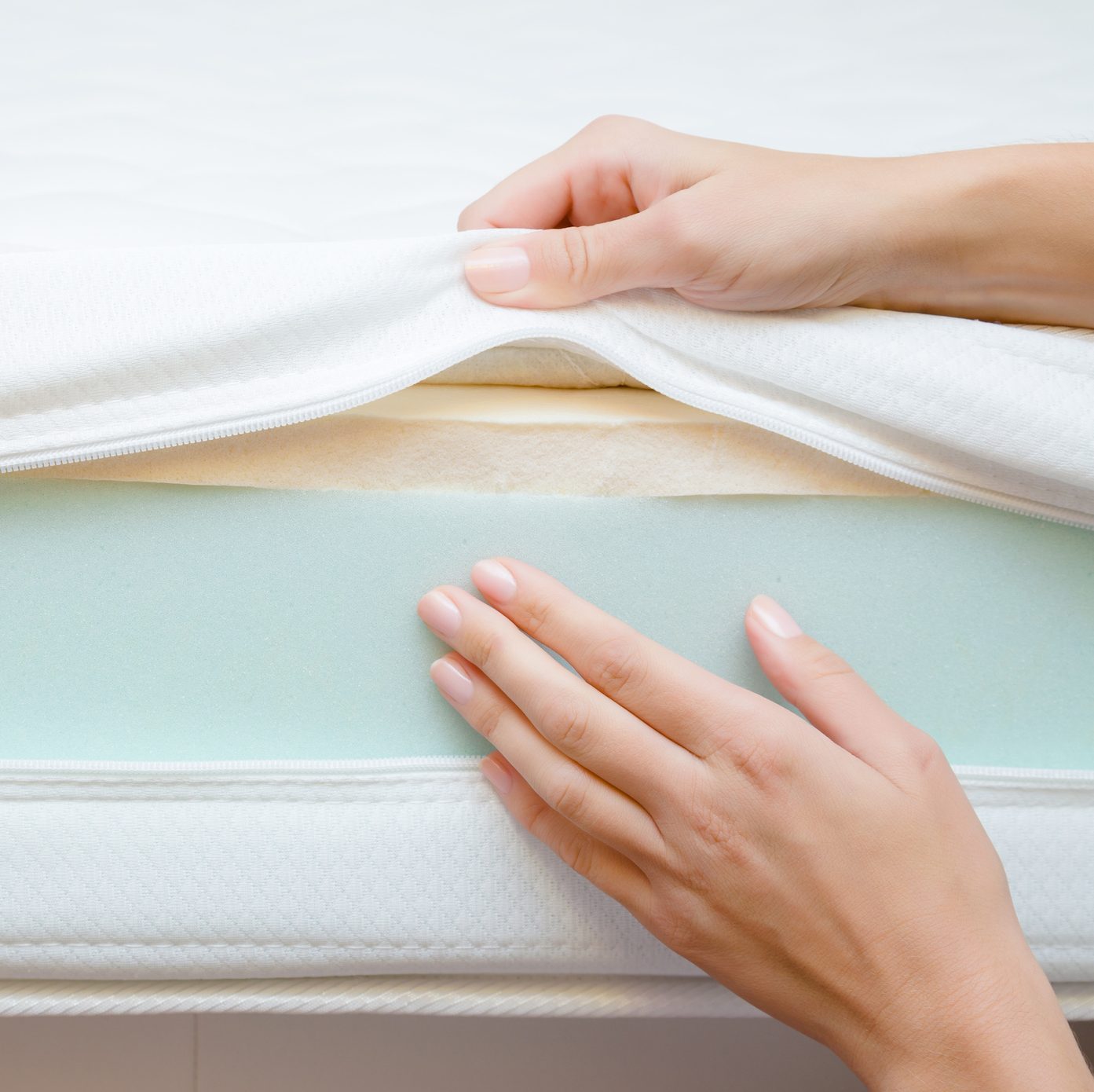 Hands unzip a mattress cover, revealing layers of foam, including a green layer, on a light-colored surface. The environment is bright and minimalistic.