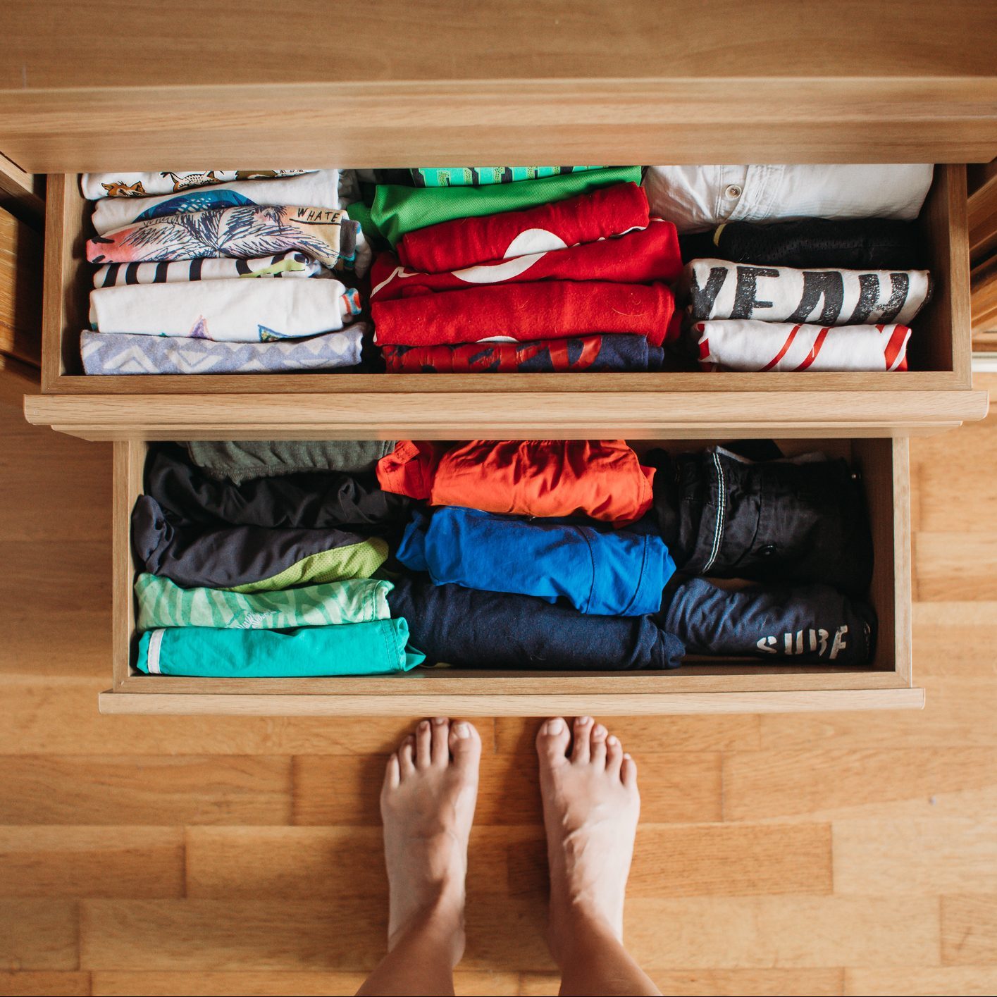 A person stands barefoot in front of two open wooden drawers filled with neatly folded colorful clothing, arranged by color and type.