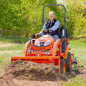 A man operates a Kubota tractor, using its plow attachment to till soil in a grassy field surrounded by trees under a clear sky.