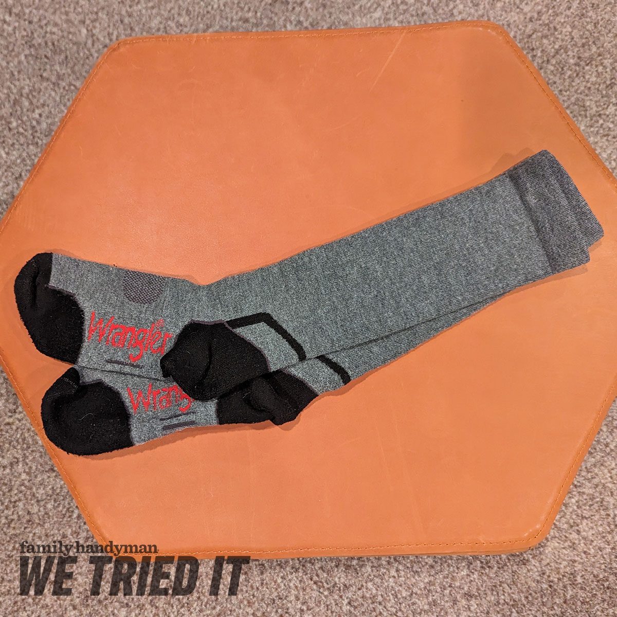 Gray socks with black accents rest on an orange hexagonal surface, showcasing the brand name "Wrangler" in red on the toes.