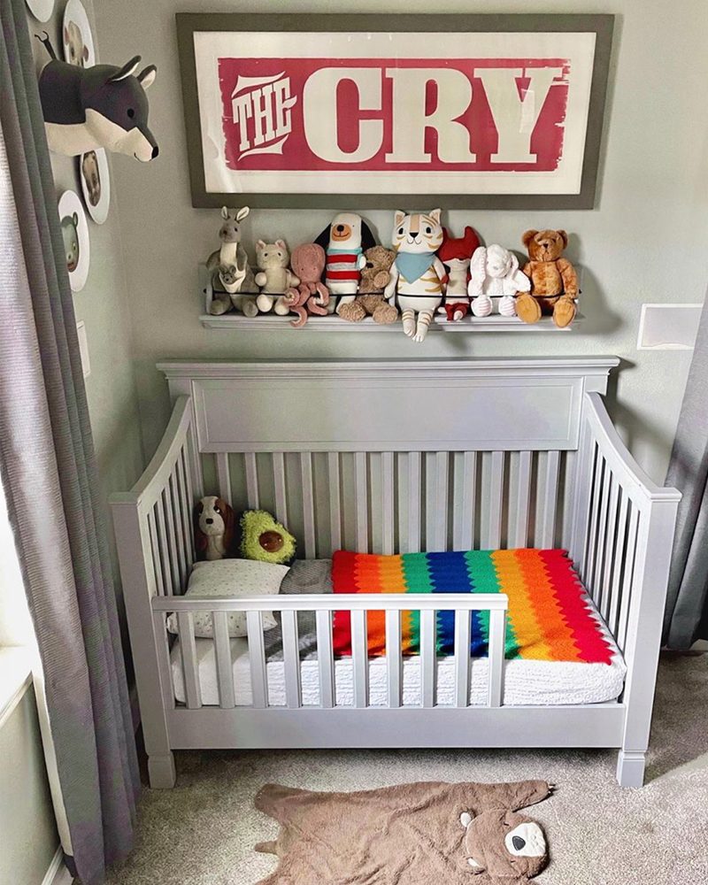 A gray crib holds colorful blankets and stuffed toys, surrounded by playful decorations on the walls, in a cozy nursery with soft carpet.