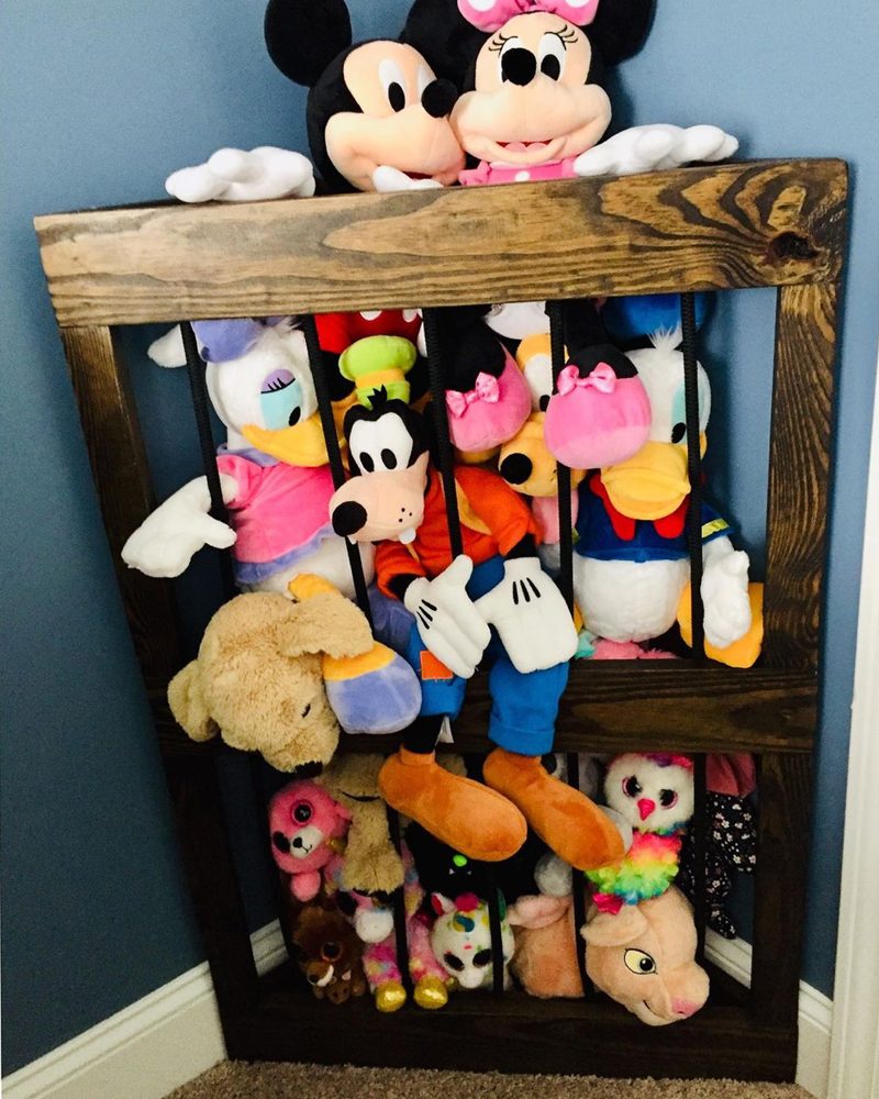 A wooden storage frame holds various plush toys, including Disney characters, arranged vertically against a blue wall and beige carpet.