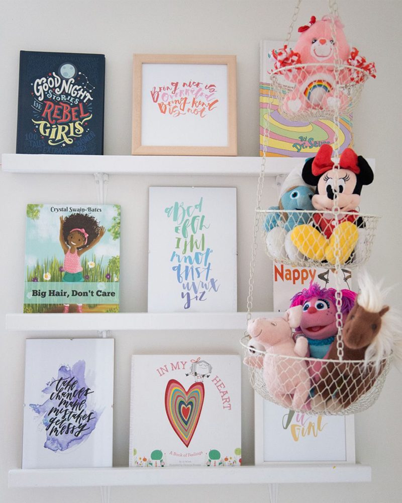 A wall shelf displays colorful children's books and framed artwork, while two hanging baskets hold plush toys, creating a playful, vibrant decor.