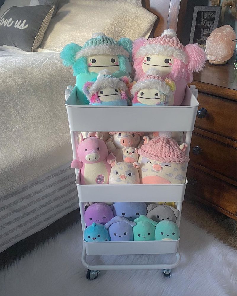 A rolling cart displays various colorful plush toys, including animal-shaped figures, stacked vertically. The cart is positioned beside a bed with soft bedding.