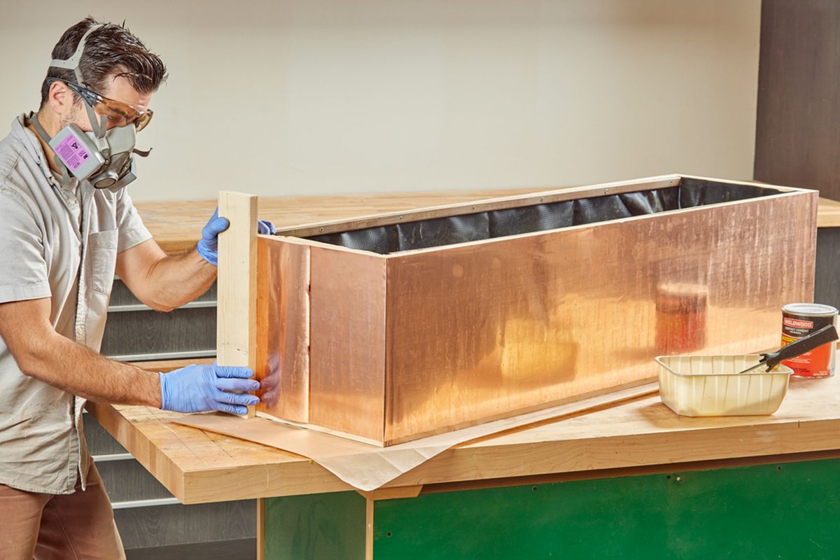 A man wearing safety gear assembles a copper planter on a wooden work surface, surrounded by tools and materials for crafting.