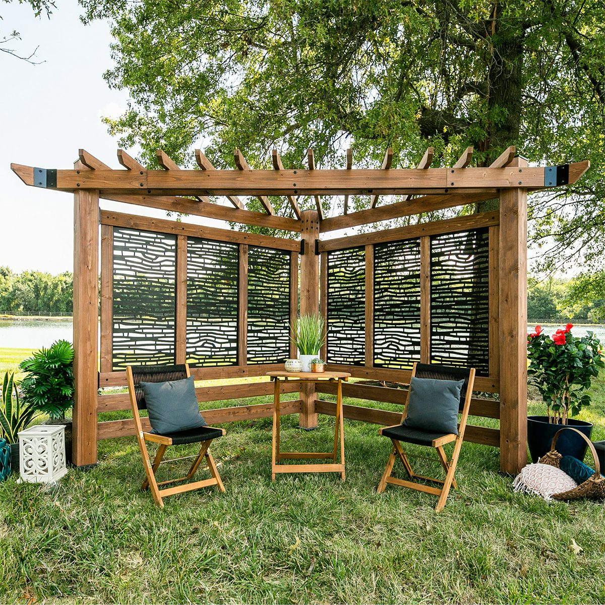 A wooden gazebo with patterned screens contains two chairs and a table, surrounded by grass, plants, and a view of a nearby body of water.