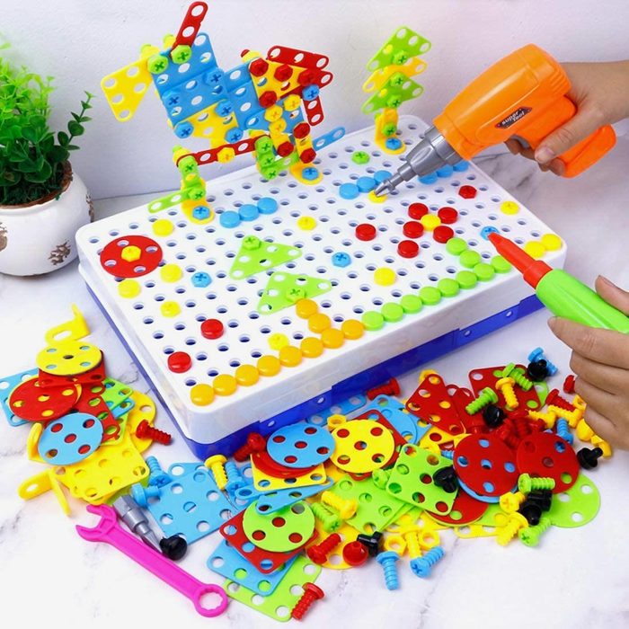 Colorful plastic building pieces are being assembled with a toy drill on a grid board, surrounded by additional pieces scattered on a surface.