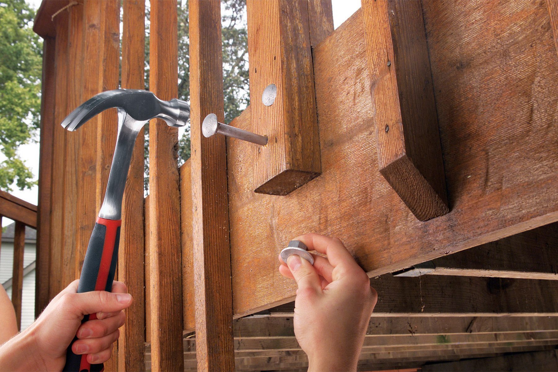 A person holds a hammer in their right hand, driving a nail into a wooden surface. Their left hand holds a large coin, presumably to protect the wood from being damaged by the hammer during the nailing process. The background shows wooden beams and outdoors.